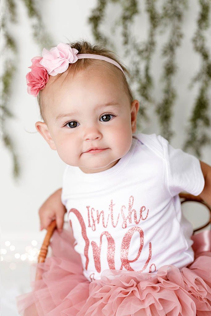 Baby girl in pink tutu and headband wearing an Isn't She One shirt celebrates her first birthday.