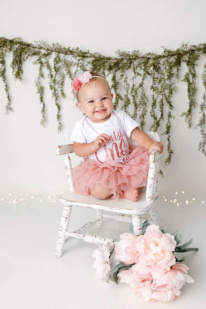 Smiling baby girl in pink tutu sits in white chair wearing ‘Wild One’ onesie with floral greenery backdrop.