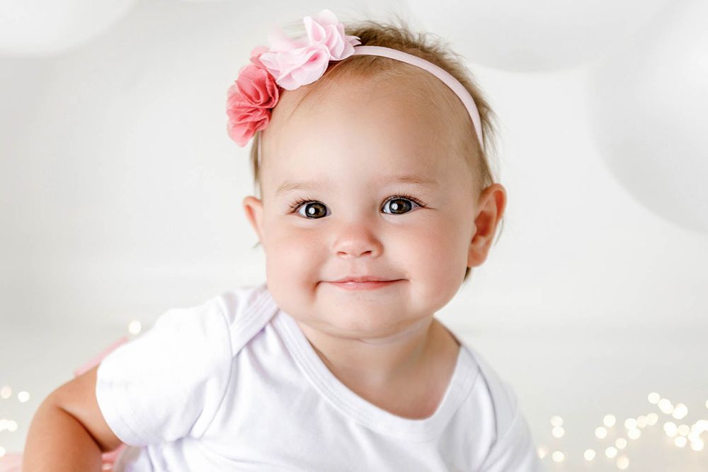 Adorable baby girl smiling wearing a white onesie and pink floral headband with bokeh light background.