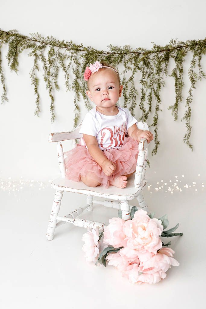 Baby girl in pink tutu sits on white chair with floral garland backdrop and pink peony flowers.
