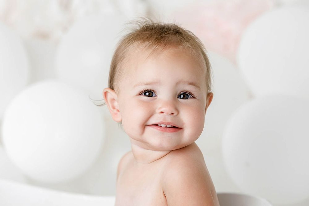 Happy baby girl smiling in white bath surrounded by white balloons by Temescal Valley cake smash photographer.