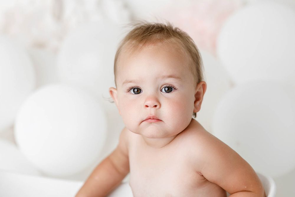 Adorable baby with big blue eyes sits surrounded by white balloons in a soft, dreamy white studio setting.
