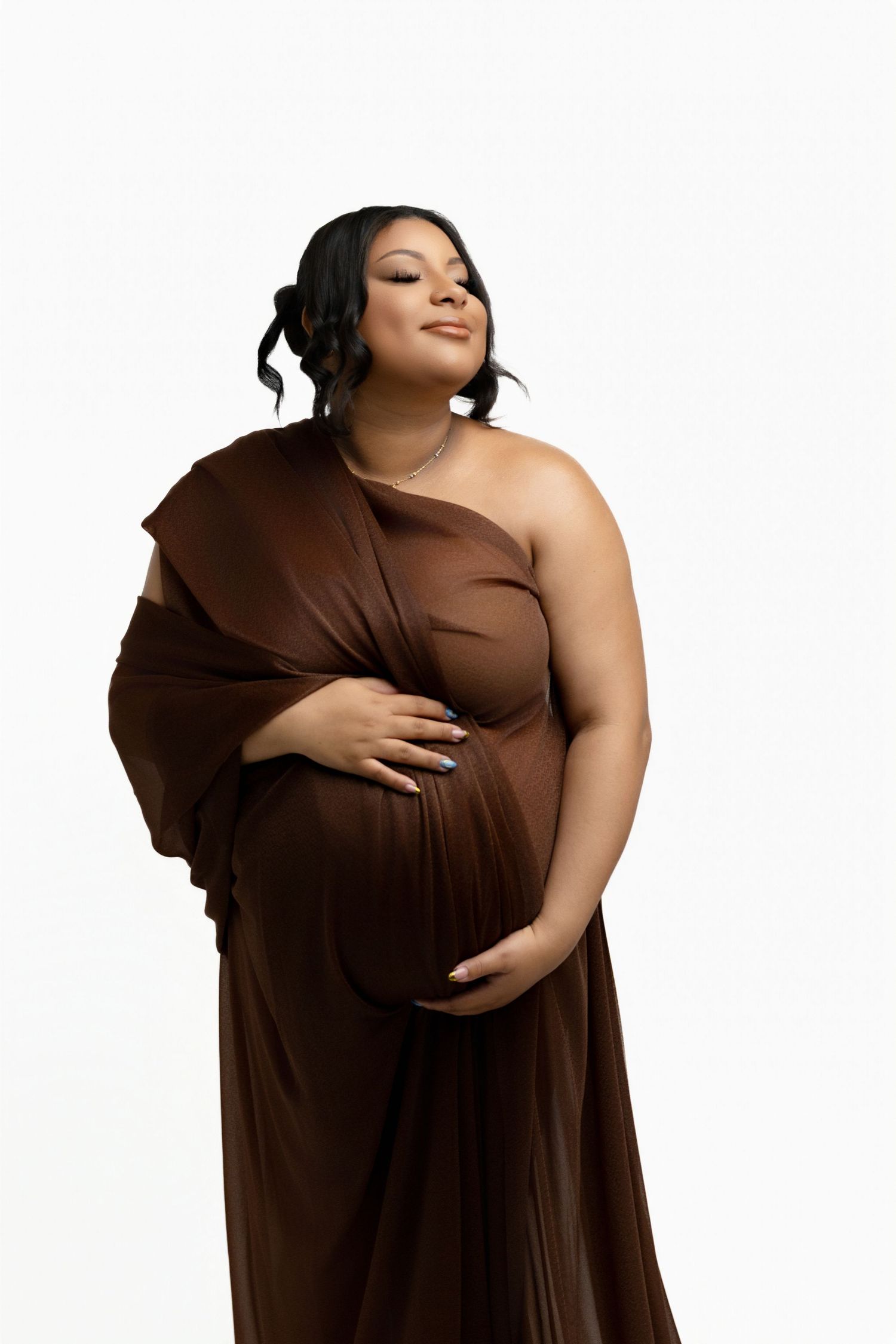 A woman with black hair smiling and looking up towards the lighting while holding her baby bump