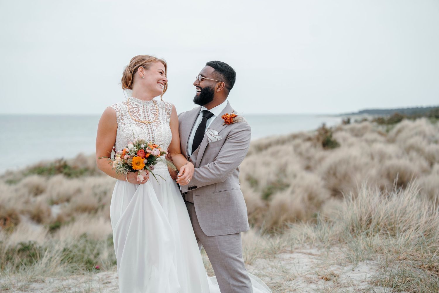 Bride and groom smiling at each other on a beach, she holds orange bouquet, he wears gray suit, ocean in background.