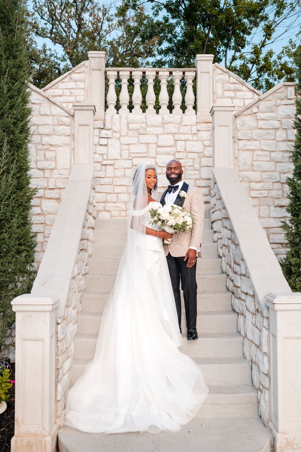 Stone staircase and white stone wall backdrop create elegant setting for wedding portraits in an outdoor garden.