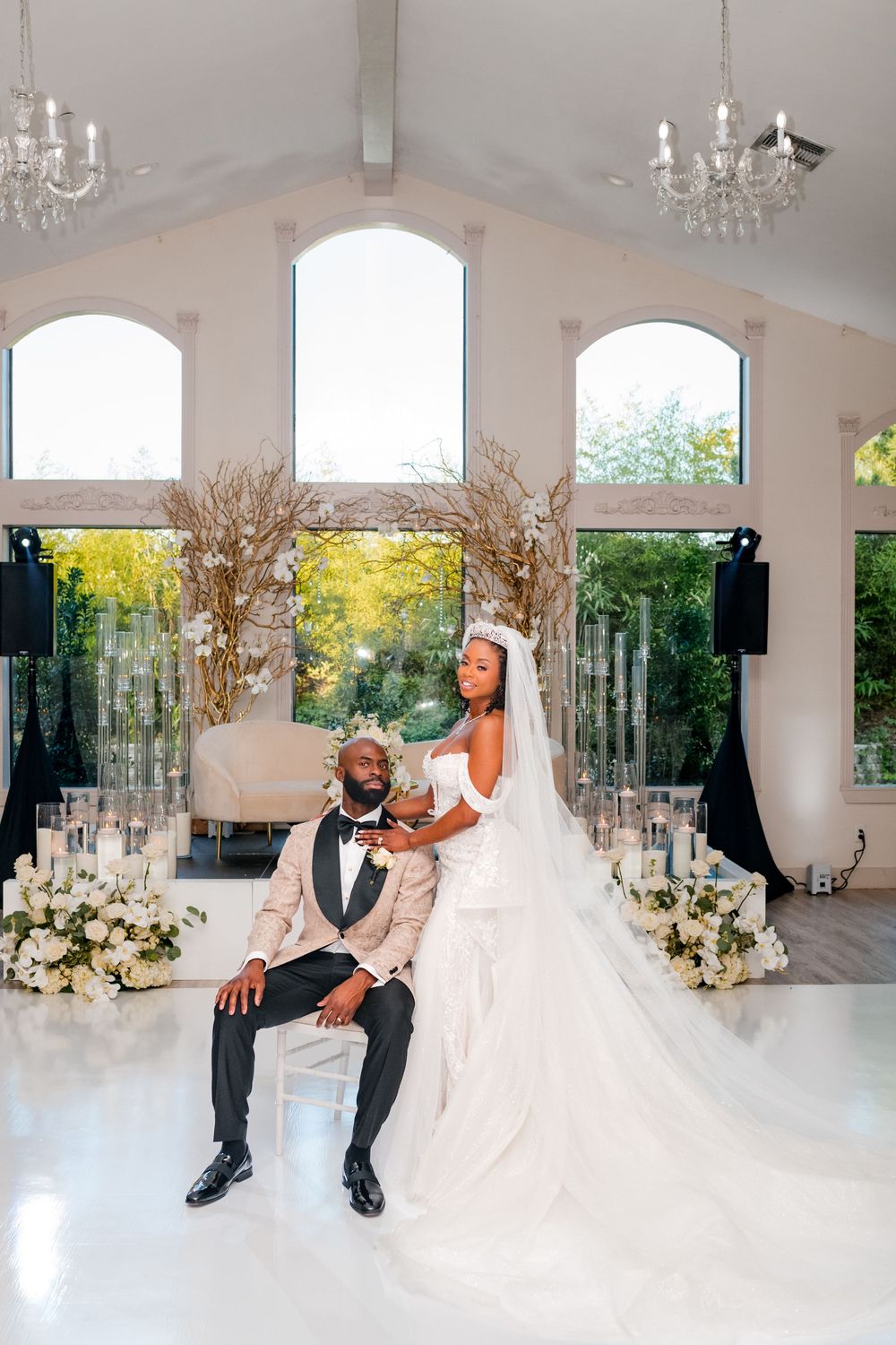 A couple in wedding attire share a romantic moment in a bright reception hall decorated with crystal chandeliers and floral decor.