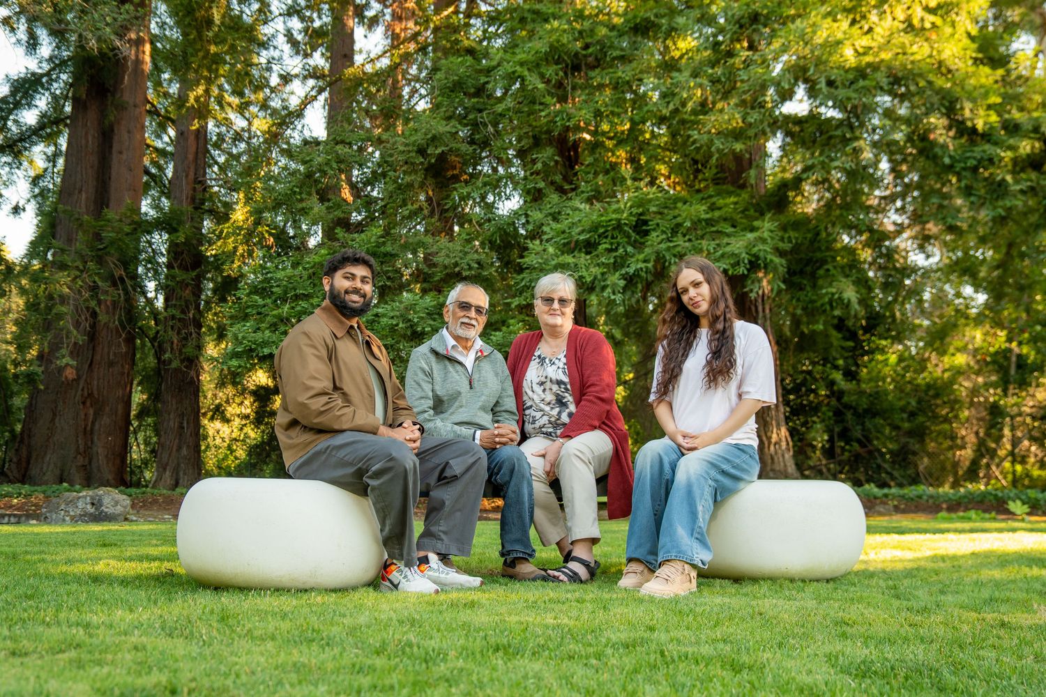 Four people sitting on white round seats on green grass with tall trees in background.