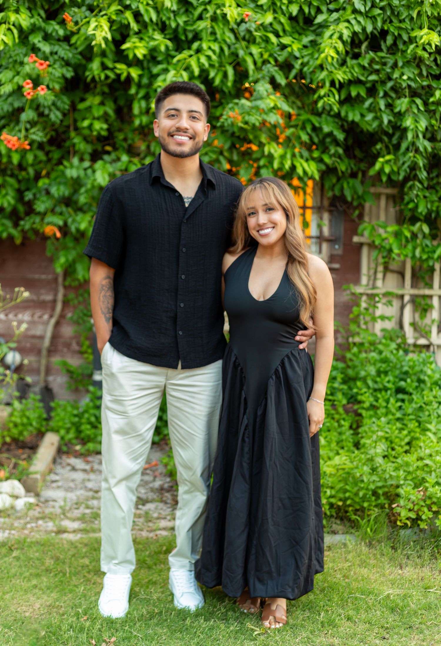 A smiling couple in matching black outfits poses together in a lush green garden setting.