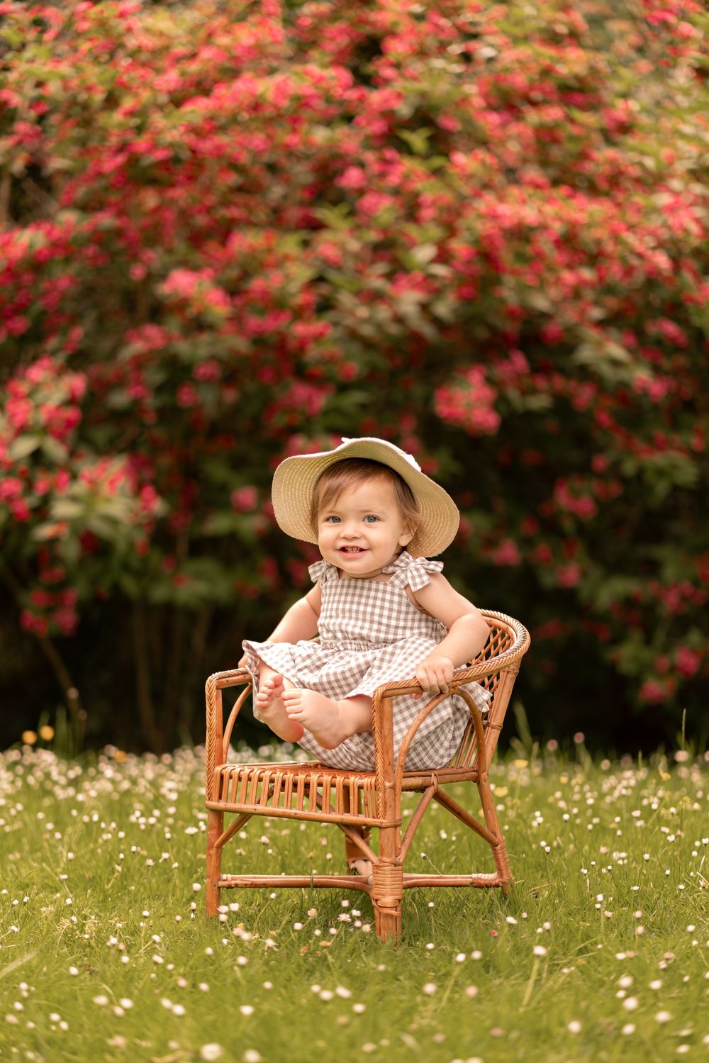 A smiling baby girl in a plaid dress and straw hat sits in a wicker chair on green grass with red flowering bushes behind.