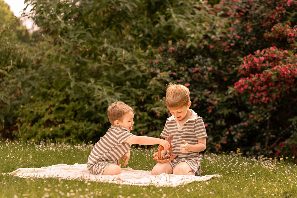 Two young boys in matching striped outfits share a playful moment on a blanket in a lush green garden.