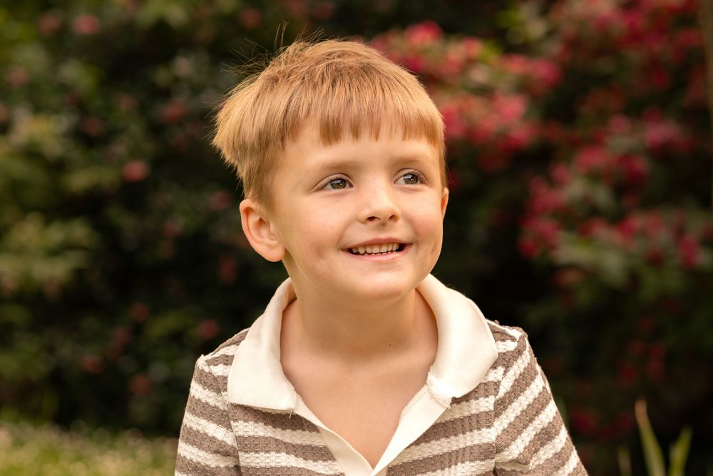 Smiling young boy with blonde hair wearing striped shirt outdoors with red flowers behind him.