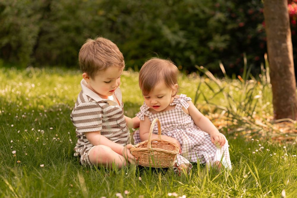 Two young children sitting on grass sharing a wicker basket in a sunny garden setting.