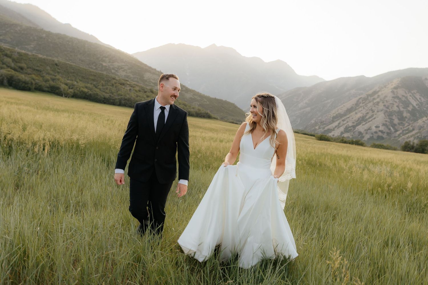 Bride and groom walking hand in hand through a lush green field with stunning mountain backdrop at golden hour.