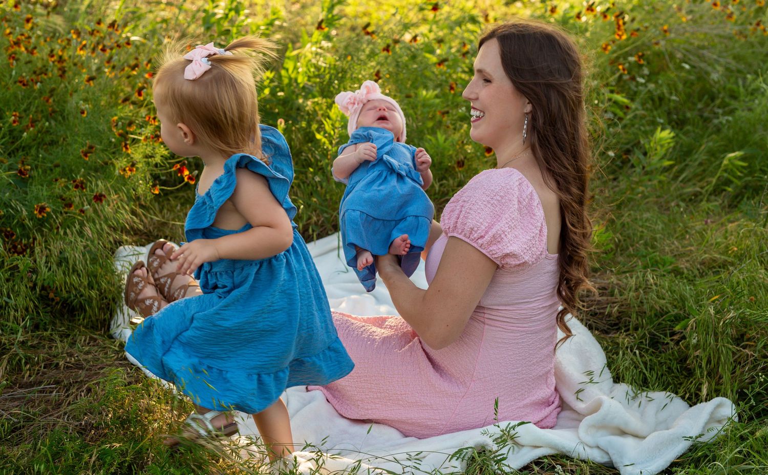 Mother in pink dress sits outdoors holding baby while toddler girl in blue dress plays in flower field.