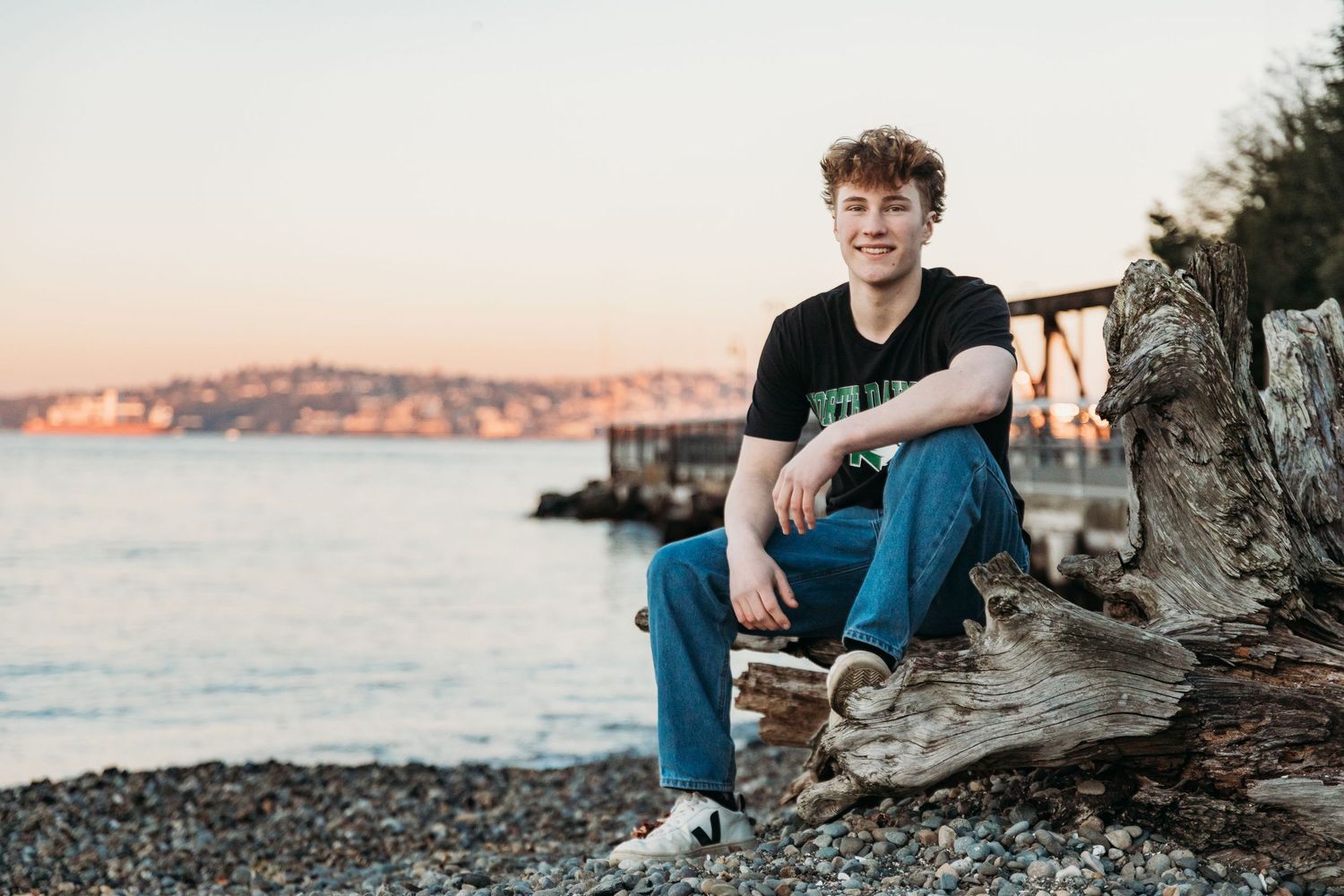 A smiling young man sits on driftwood at a rocky beach during golden hour sunset with water and city in background.