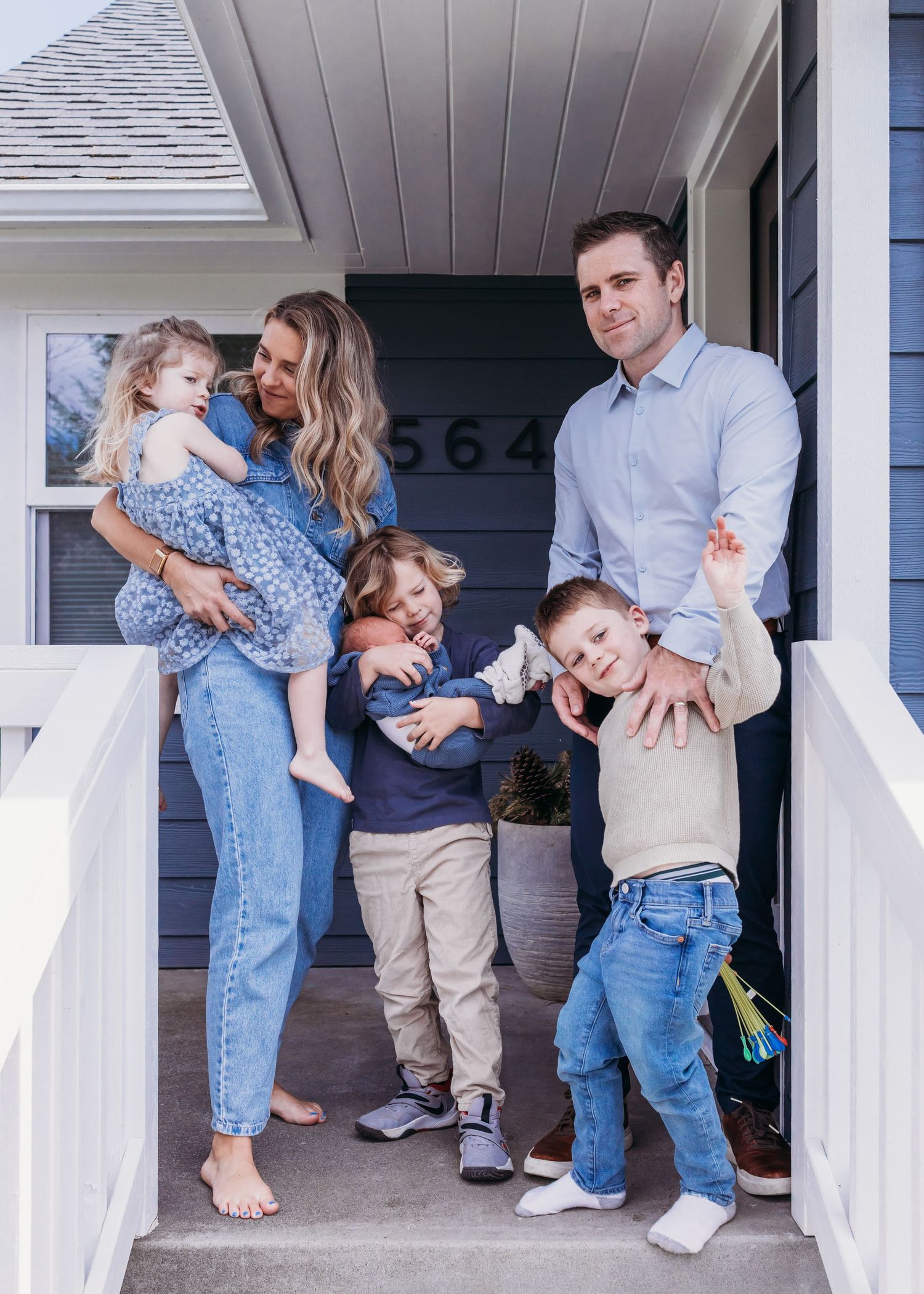 Happy family of five posing on their front porch, parents with three young children smiling outside their blue home.