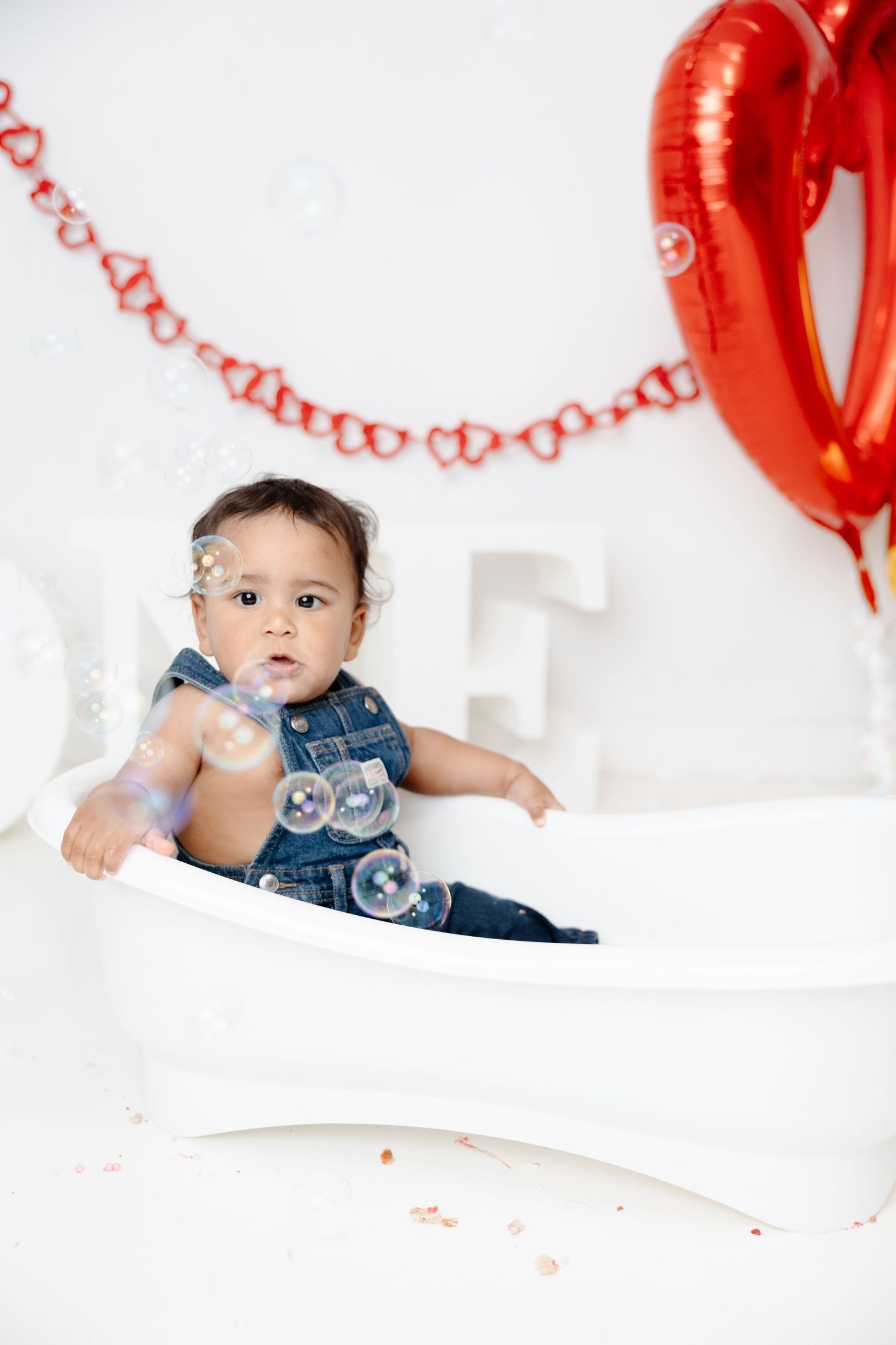 Baby in denim overalls sits in white tub with bubbles, red heart balloons and garland decorating the background.