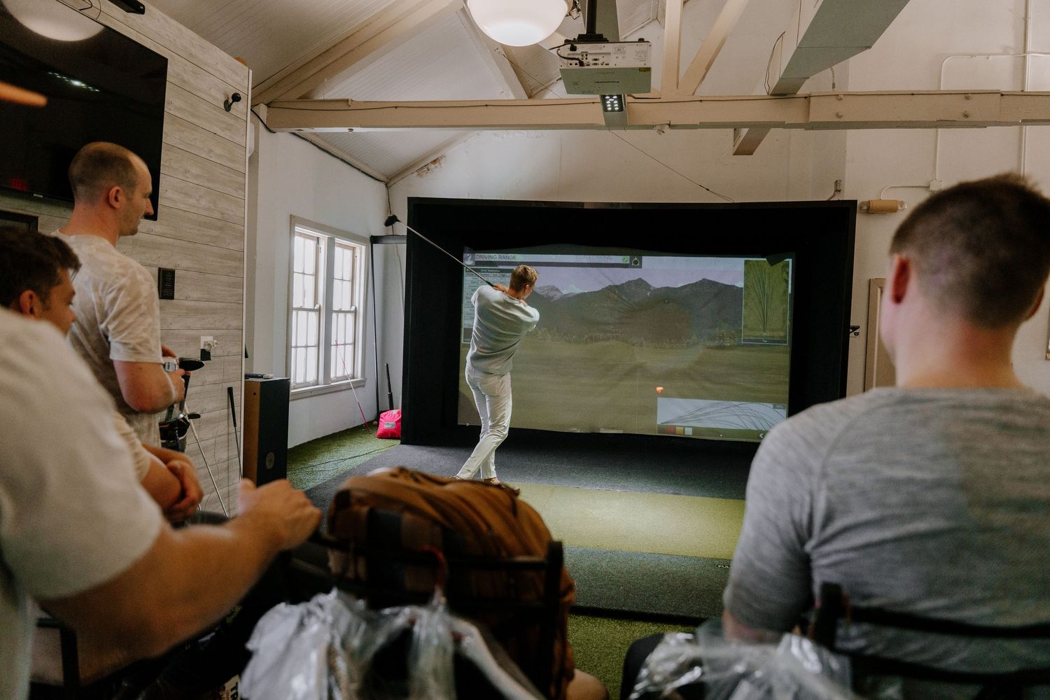 People watching a golf simulator screen in an indoor simulator bay with a golfer taking a swing.