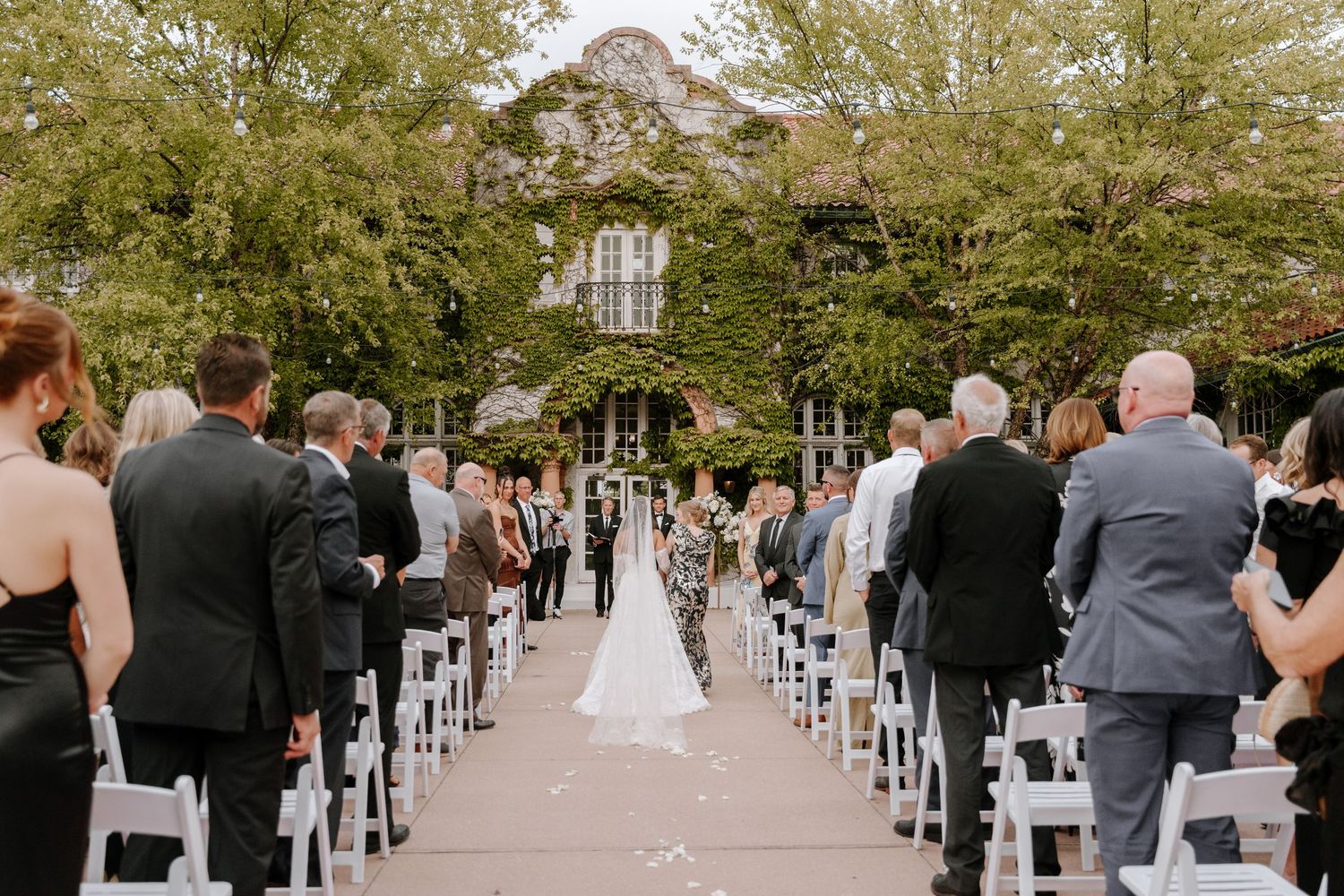 Outdoor wedding ceremony with guests seated in white chairs along an aisle leading to couple at altar near ivy-covered venue.