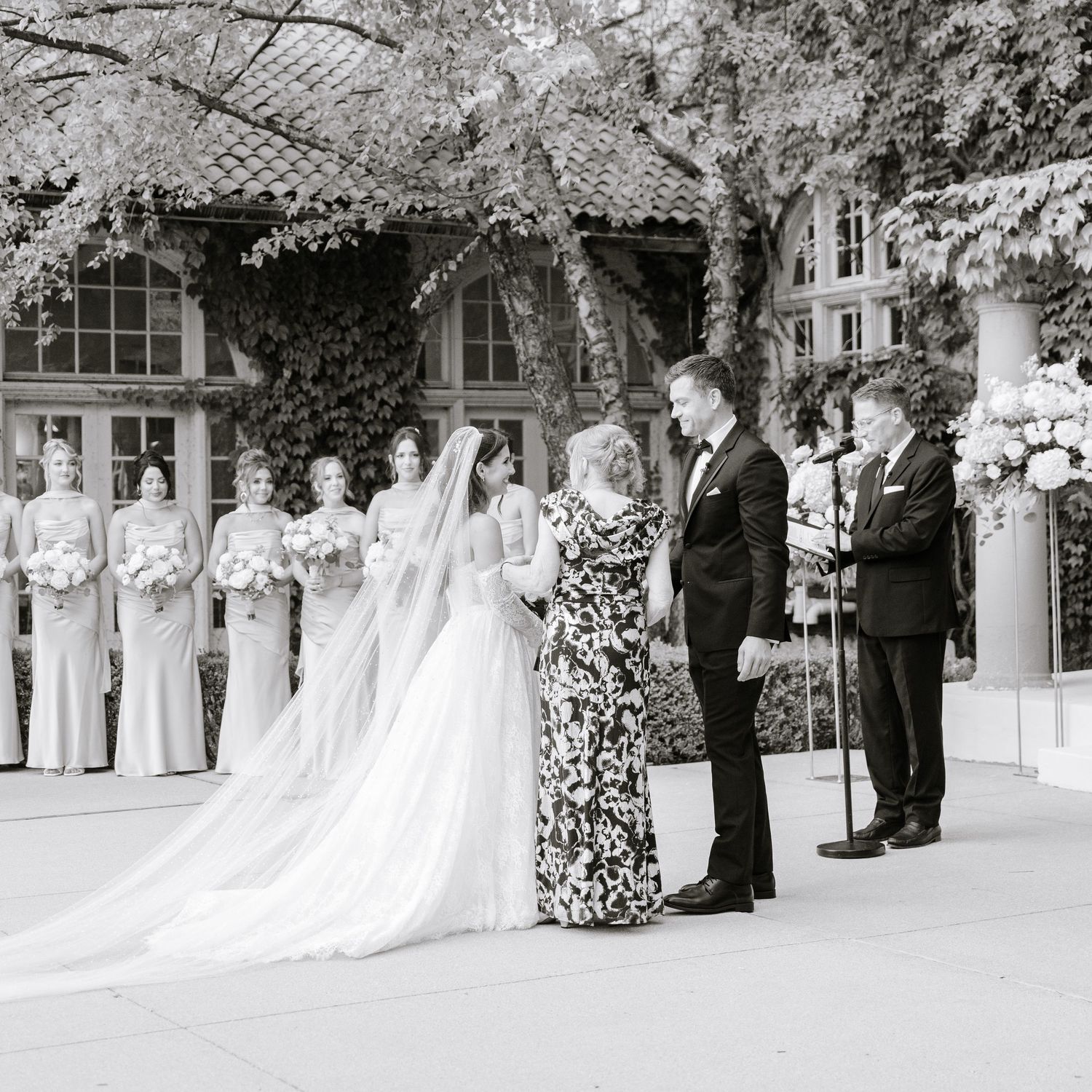 Black and white photo of bride and groom exchanging vows outdoors with bridesmaids and floral arrangements.