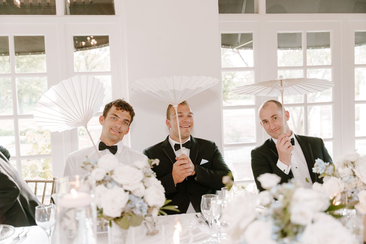 Three groomsmen in tuxedos holding white paper fans at a wedding reception table with floral centerpieces.