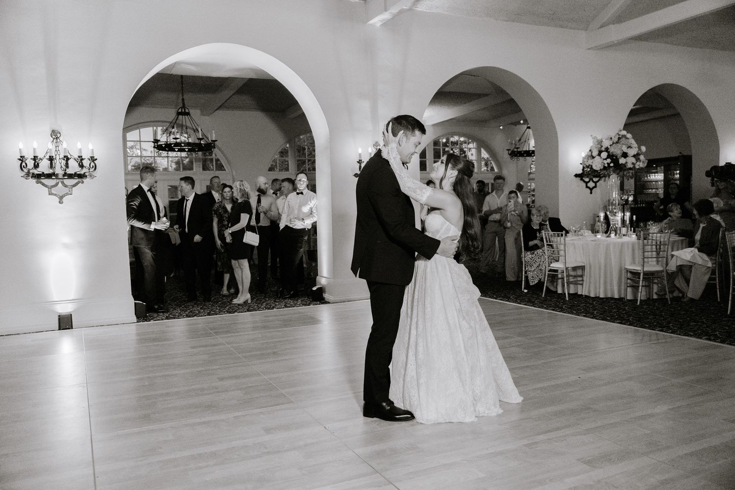 Bride and groom share their first dance in elegant ballroom while wedding guests watch in black and white photo.