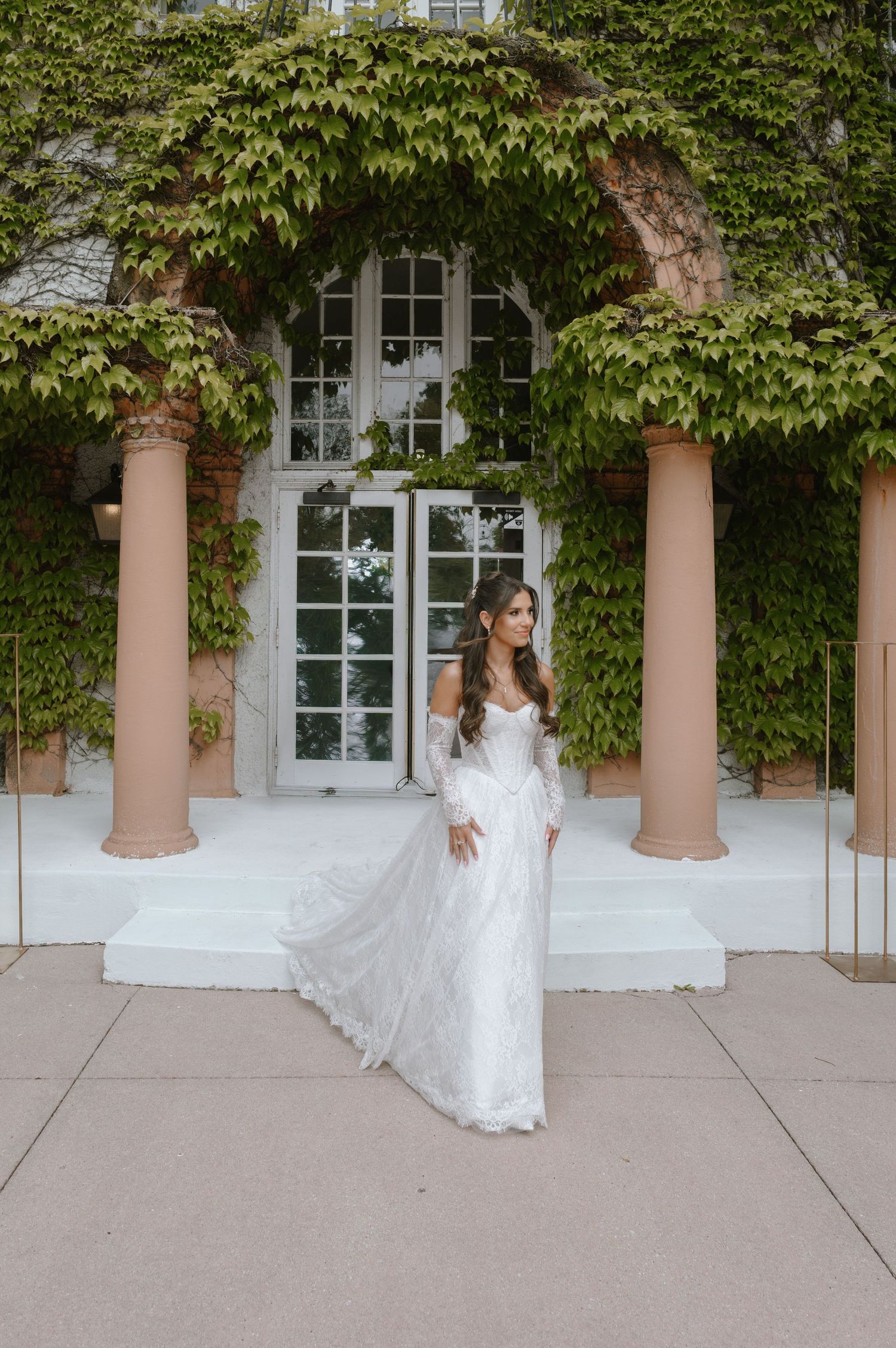 Bride in white lace off-shoulder gown with long sleeves poses before ivy-covered building with elegant columns.