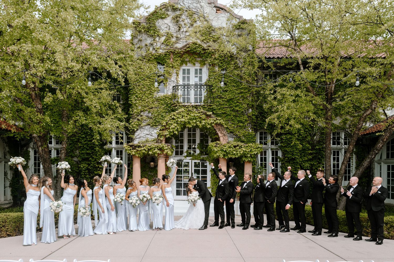 Wedding party poses outside ivy-covered venue, bridesmaids in blue gowns and groomsmen in black suits holding bouquets.
