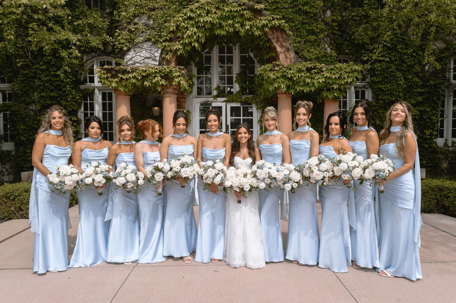 Bridesmaids in light blue satin gowns holding white bouquets stand with bride in white dress before ivy-covered building.
