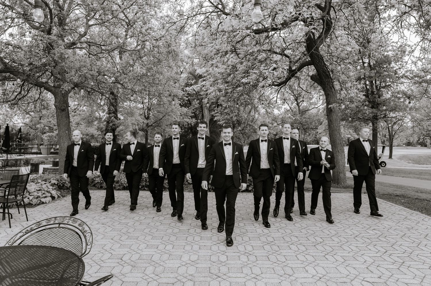 Black and white photo of a groom and groomsmen in tuxedos walking confidently outdoors among trees.