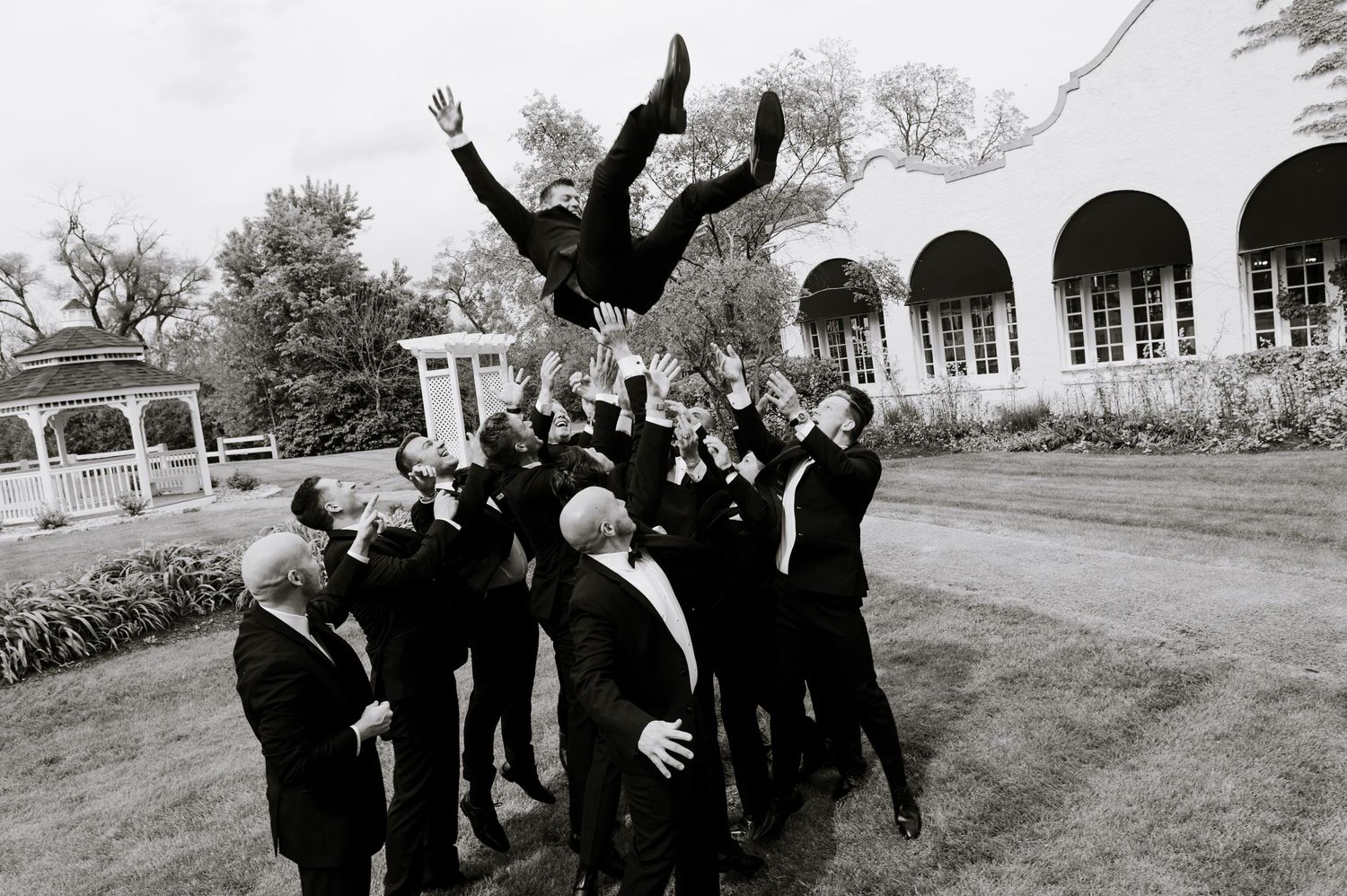 Groomsmen in black tuxedos tossing groom into the air outdoors at elegant wedding venue.