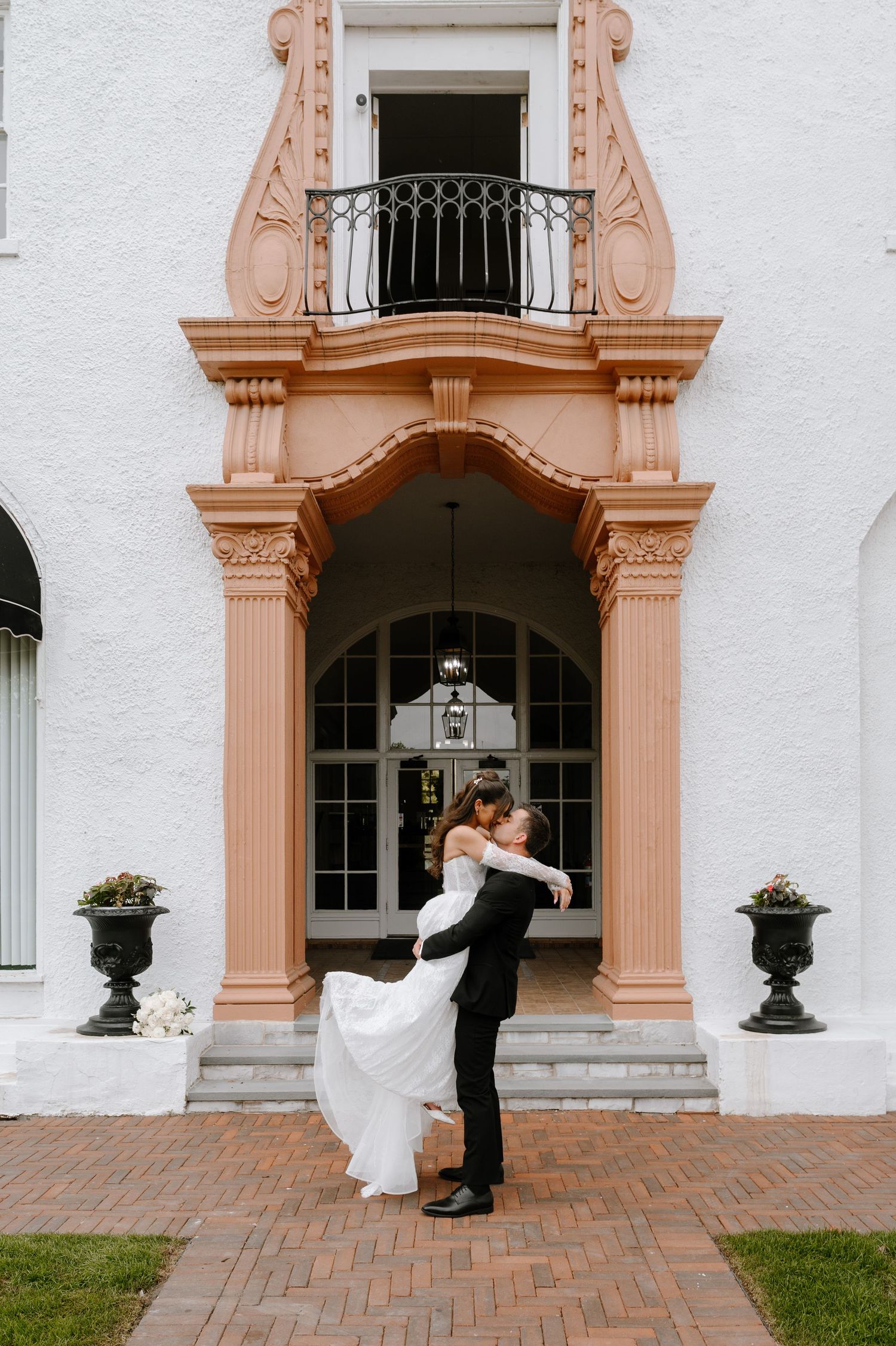 Groom lifts bride in lace wedding gown before ornate pink and white arched entrance of elegant venue.