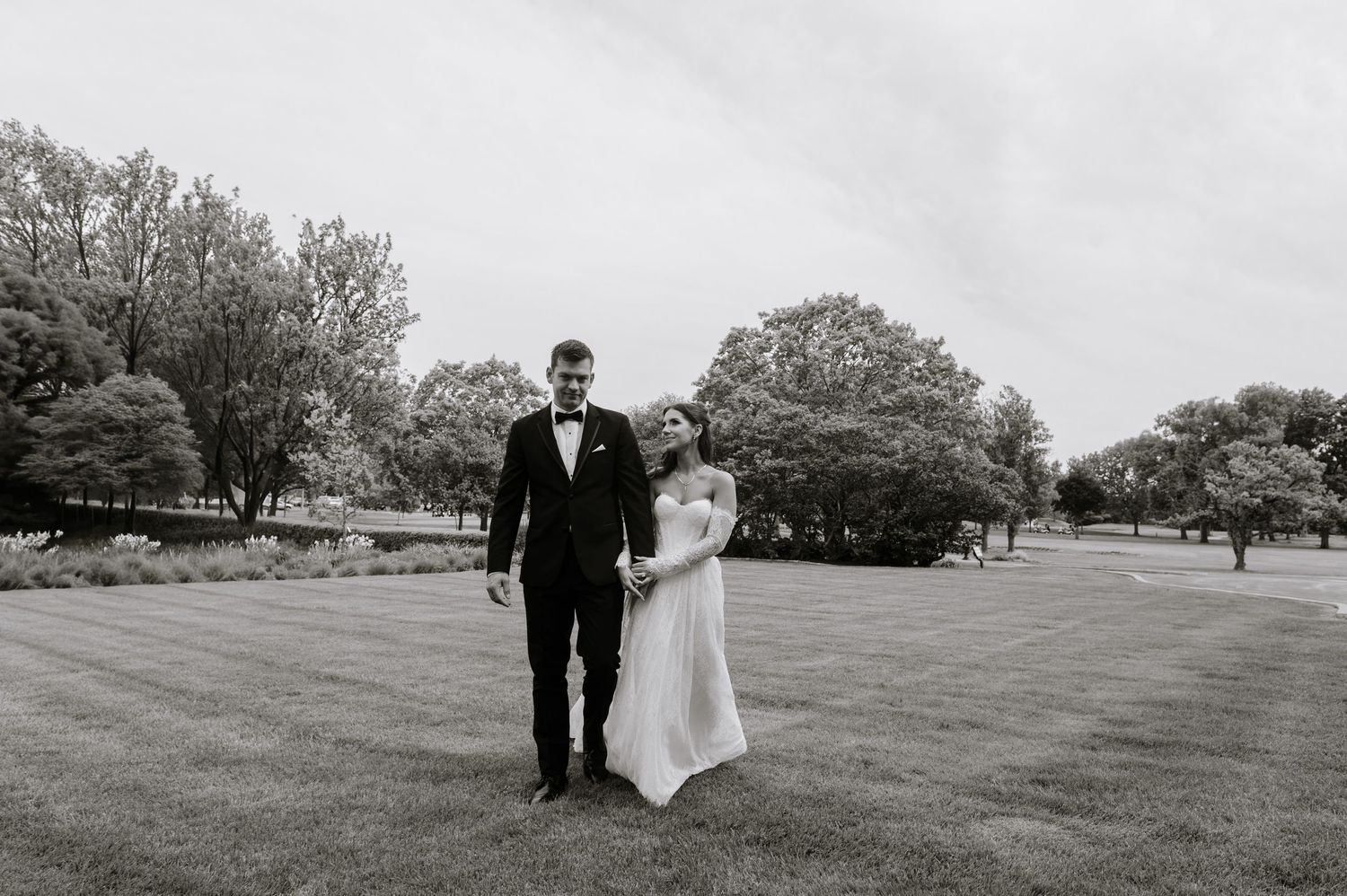 Black and white photo of a bride and groom standing on a lush green lawn surrounded by tall trees.