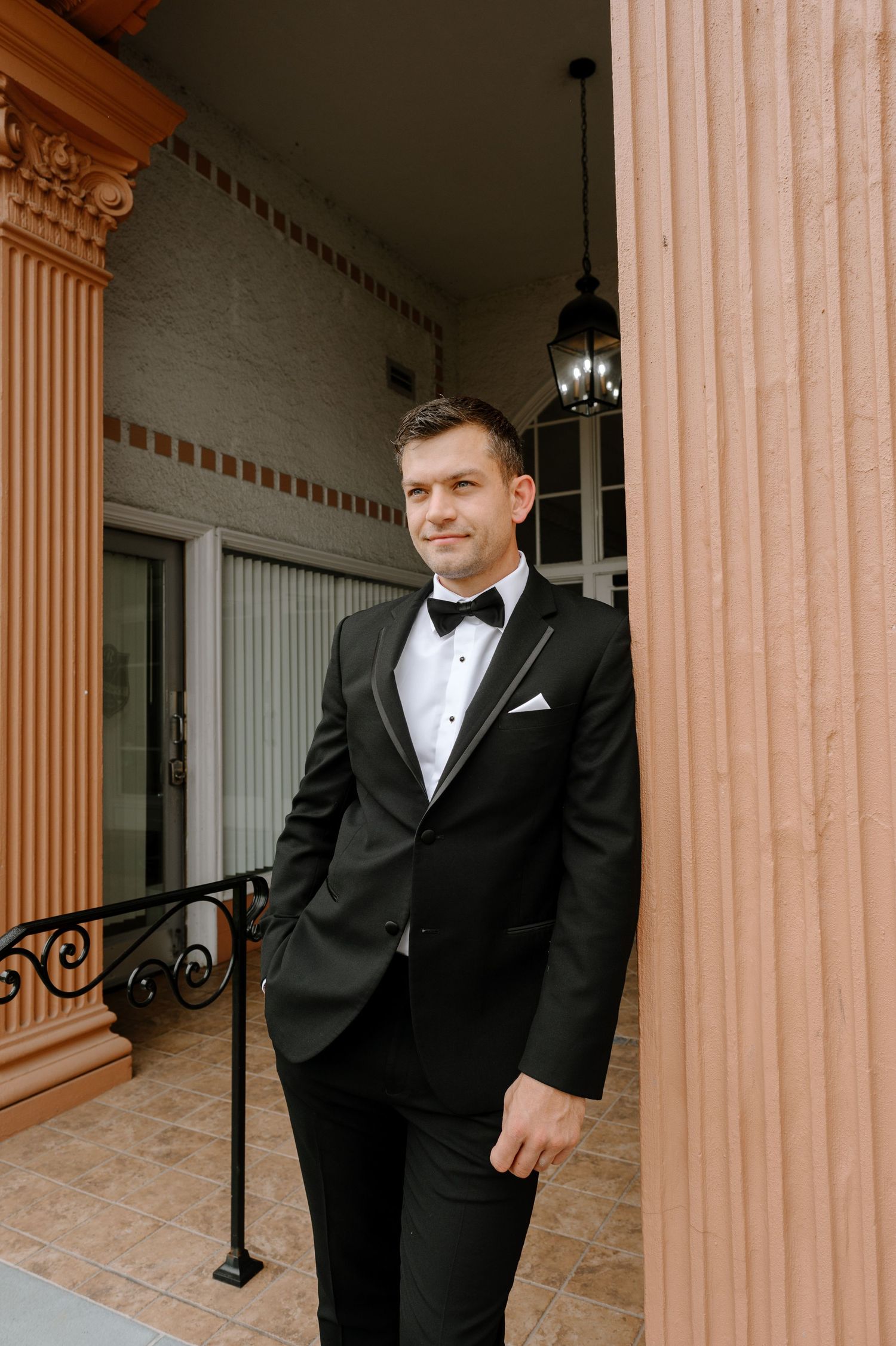 Dapper groom in black tuxedo with bow tie and white pocket square poses beside terracotta column.