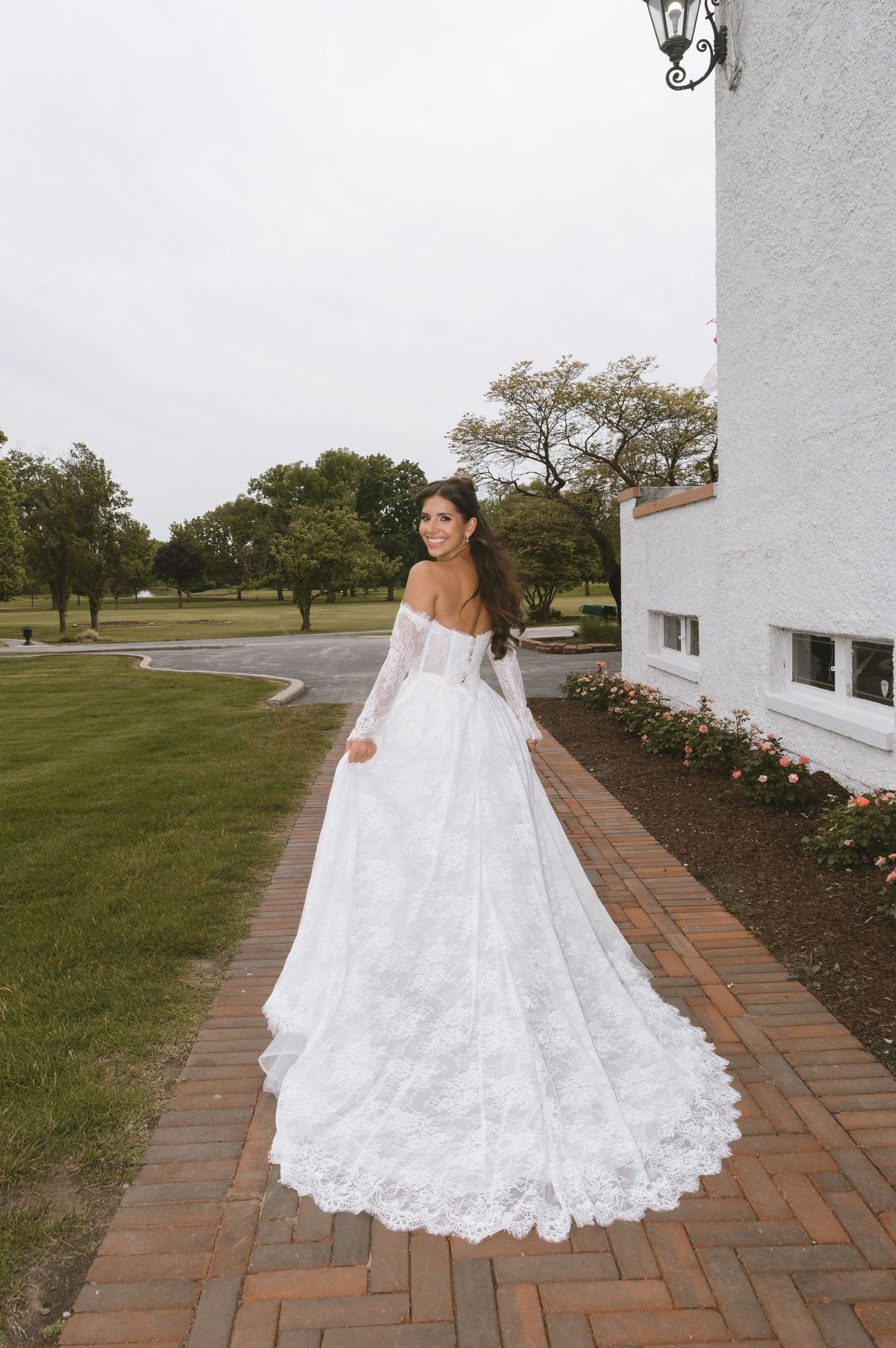 Bride in off-shoulder lace ball gown with long train walks along brick path beside white building.