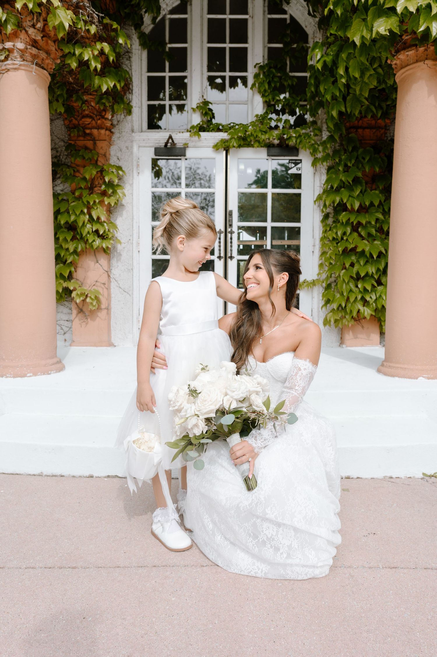 Bride in strapless lace gown kneels beside flower girl in white dress, holding bouquet outside ivy-covered building.