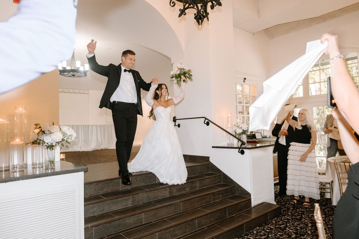 Bride and groom make grand entrance descending elegant staircase at wedding reception in white venue.