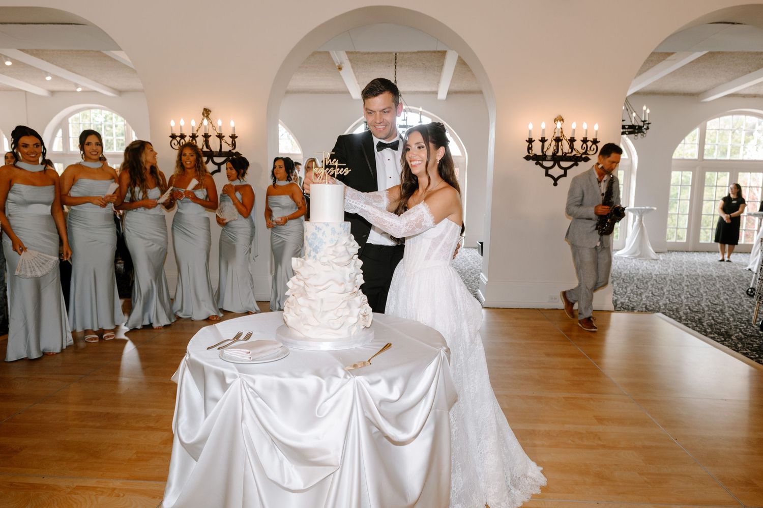 Bride and groom cut their white tiered wedding cake surrounded by bridesmaids in silver gowns in elegant venue.