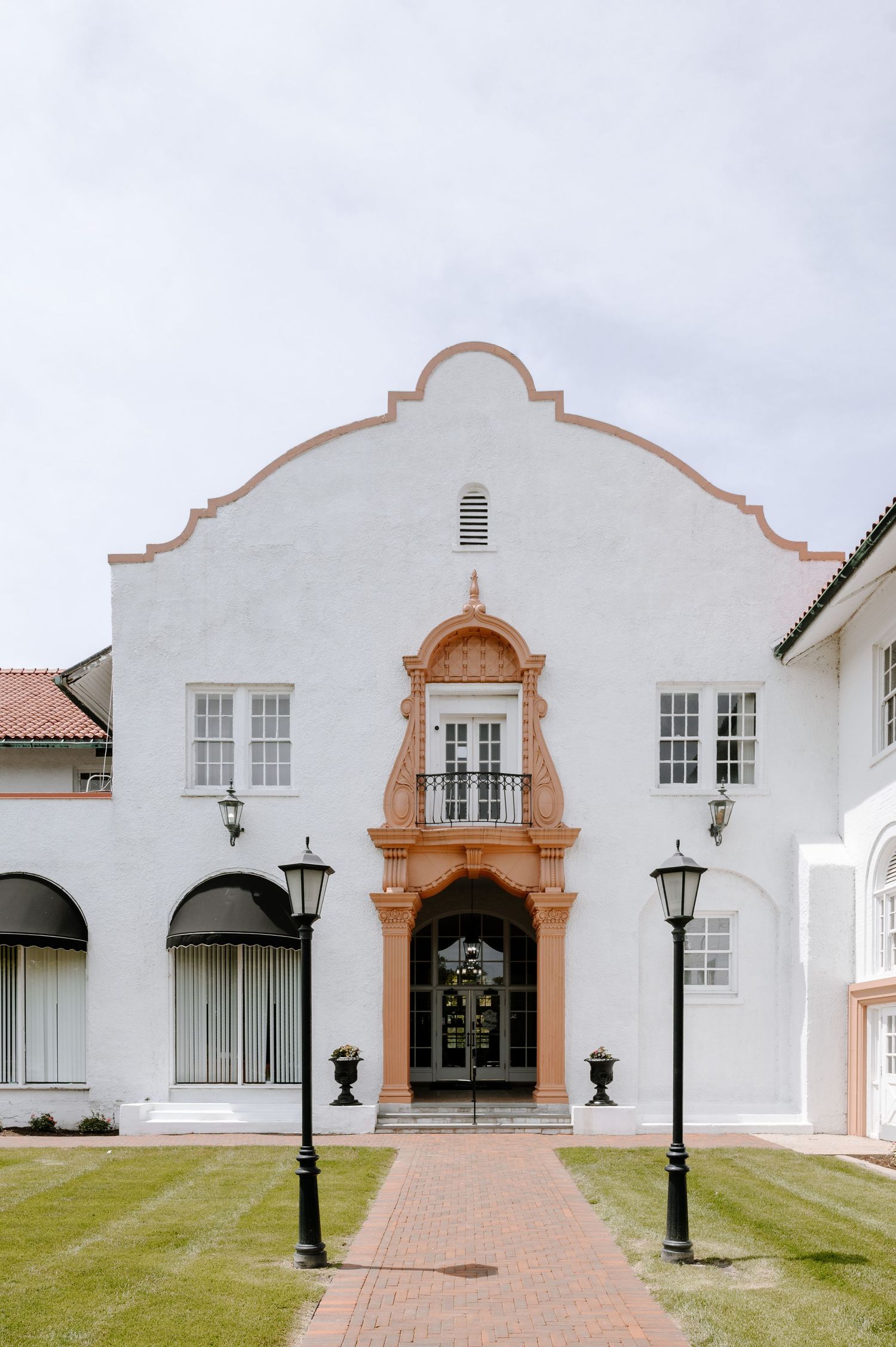 White Spanish Colonial Revival building with ornate terracotta entrance, symmetrical lamp posts, and manicured lawn under overcast sky.