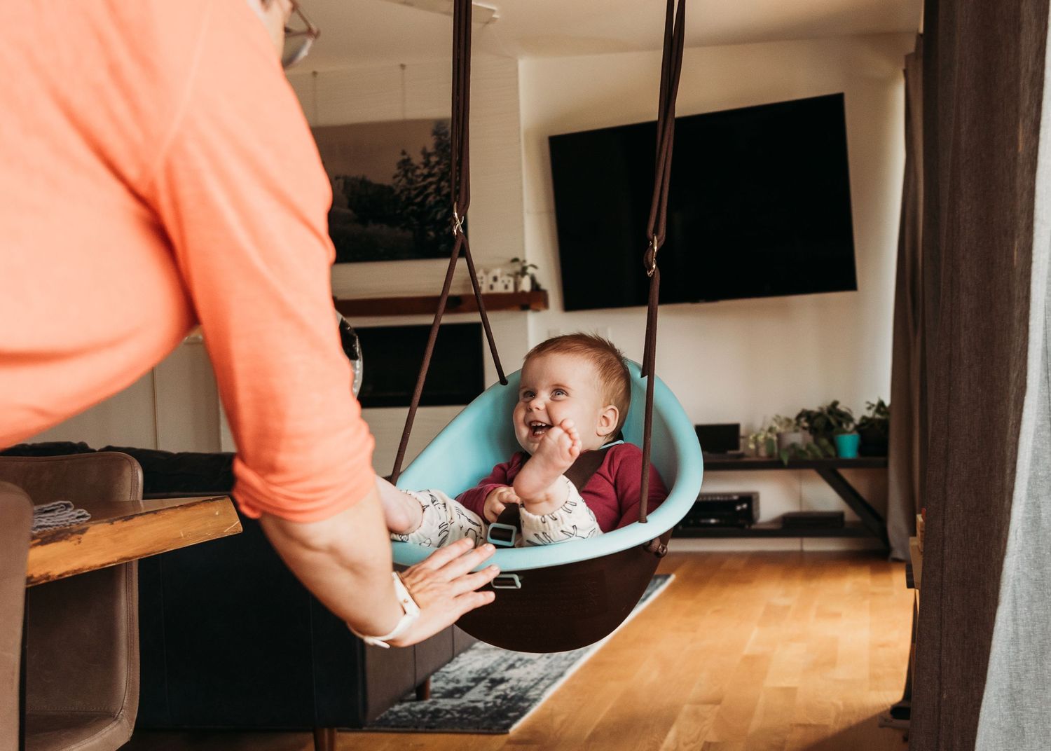 Happy baby sitting in a teal indoor swing while a parent in an orange shirt gently pushes them.