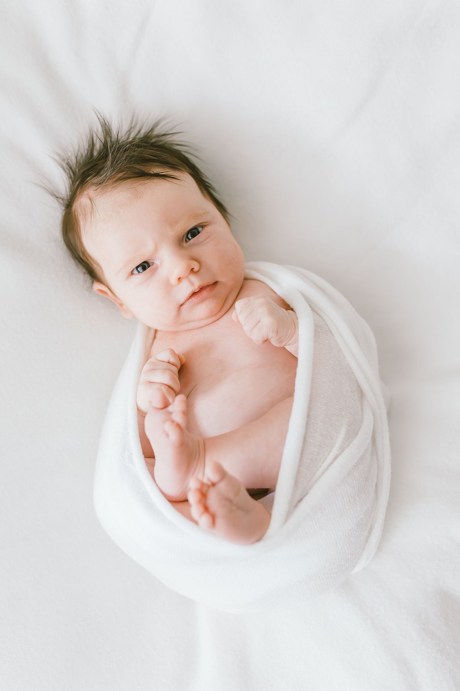 Newborn baby swaddled in white wrap lying on white blanket, with dark hair and alert eyes looking up.