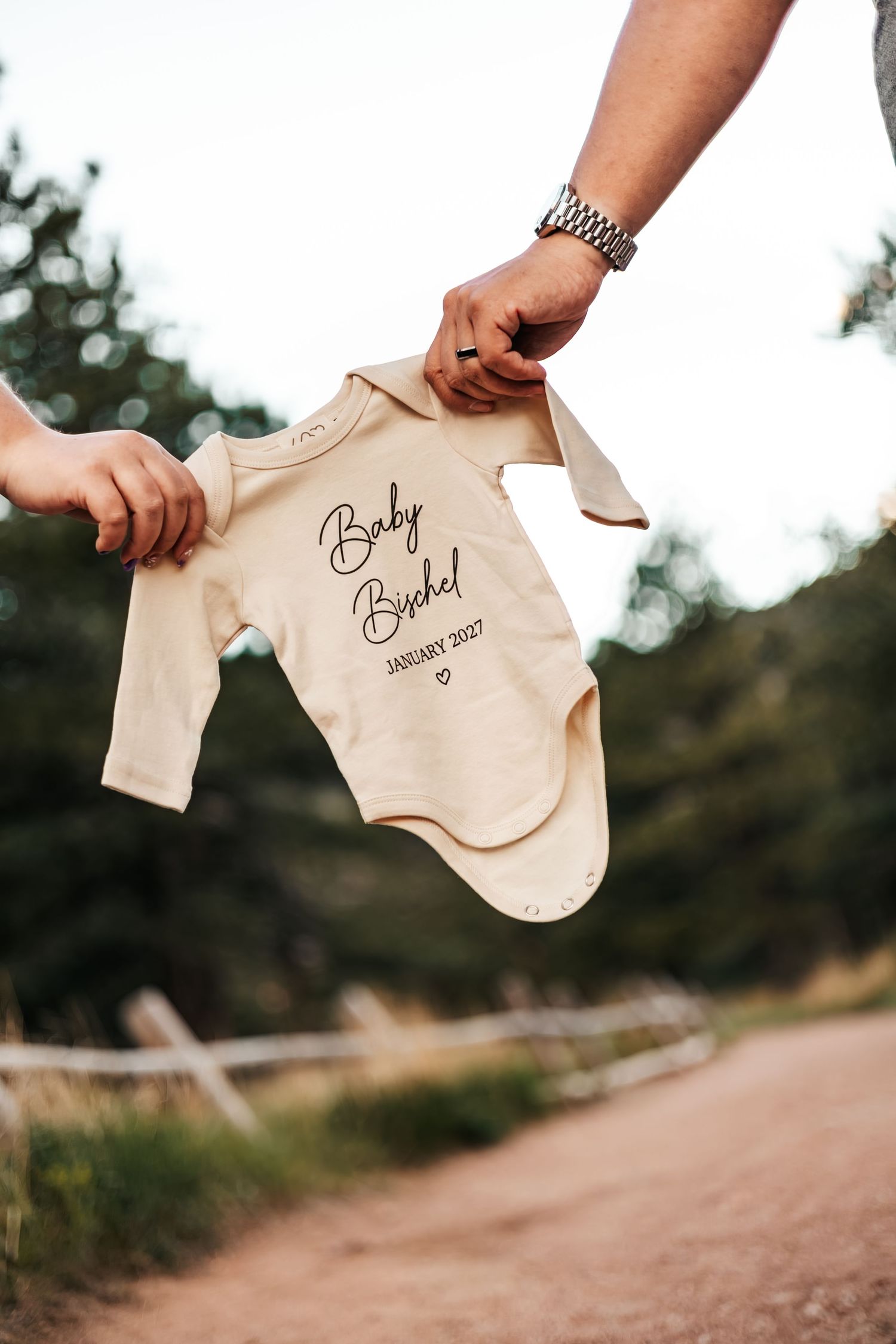 Two hands holding a cream baby onesie printed with Baby Bischel January 2027  | Baby Announcement Photographer in Boulder, Colorado