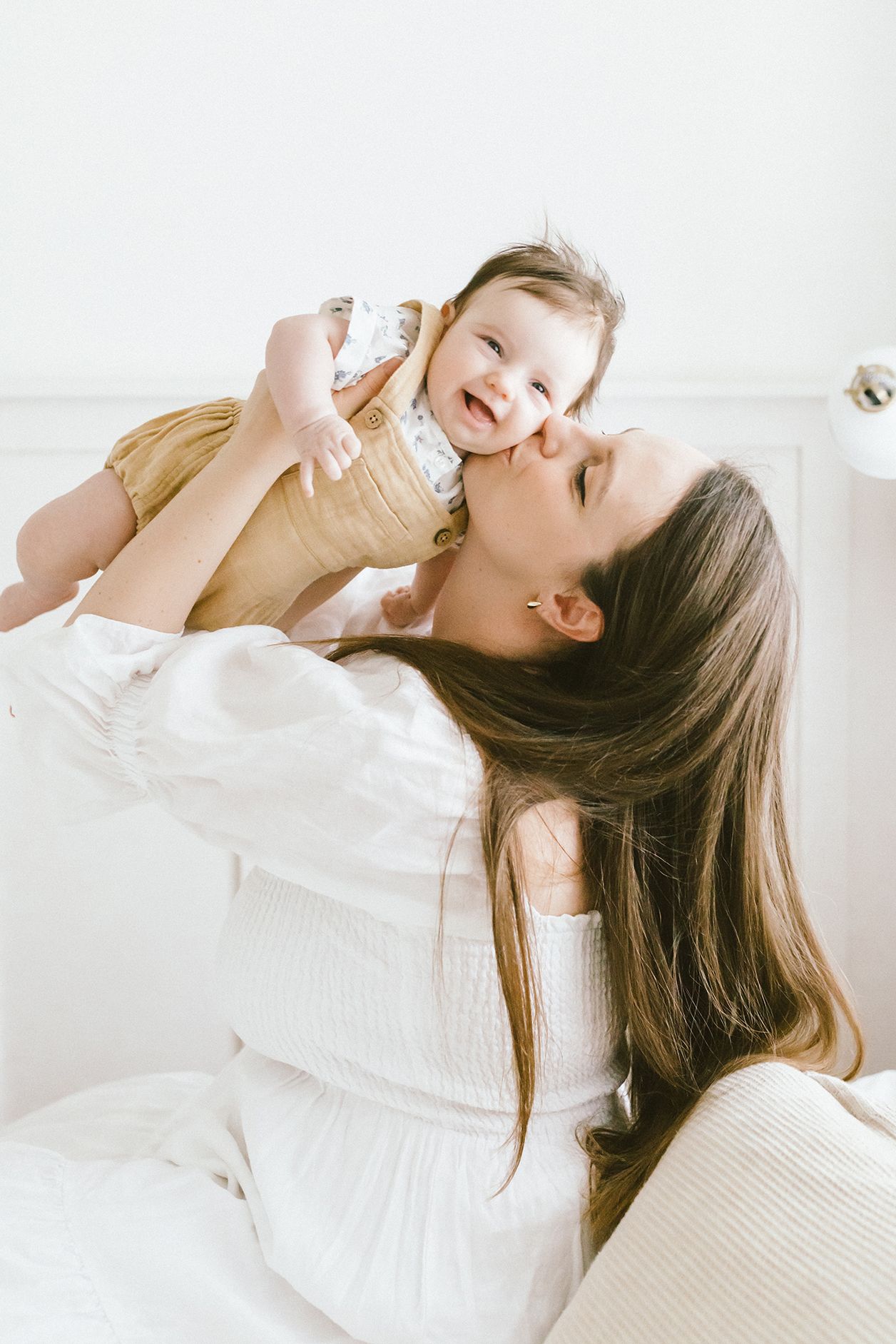 A joyful mother lifts her smiling baby girl while sitting on a white bed in a bright, airy room.