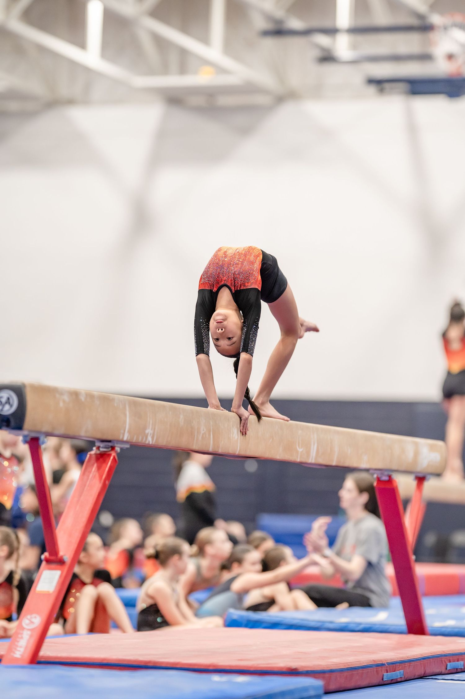 Young gymnast performing a back walkover on a balance beam during a gymnastics competition in an indoor gym.