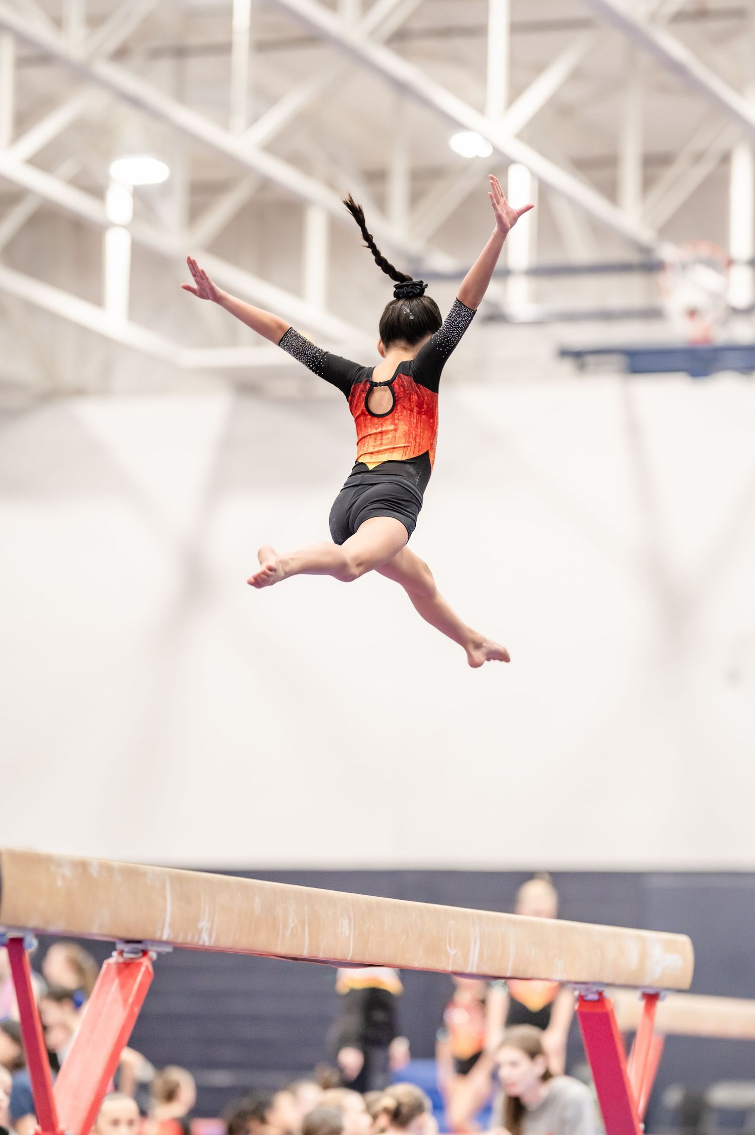 Young gymnast leaping mid-air above a balance beam in a black and red leotard during a gymnastics competition.