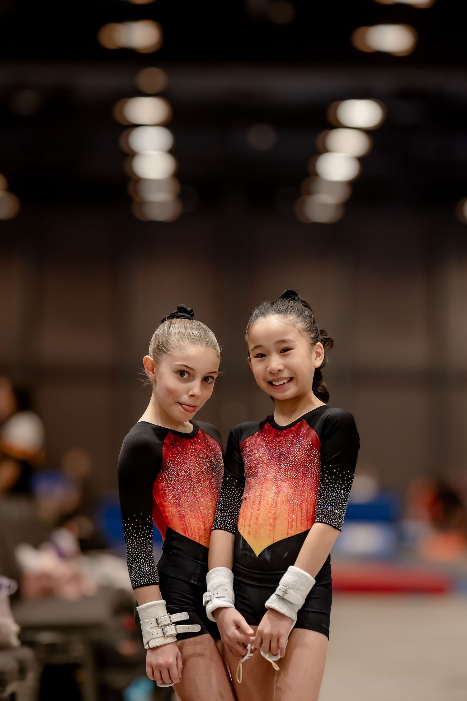 Two young gymnasts in matching black and red leotards pose together smiling at a gymnastics competition.