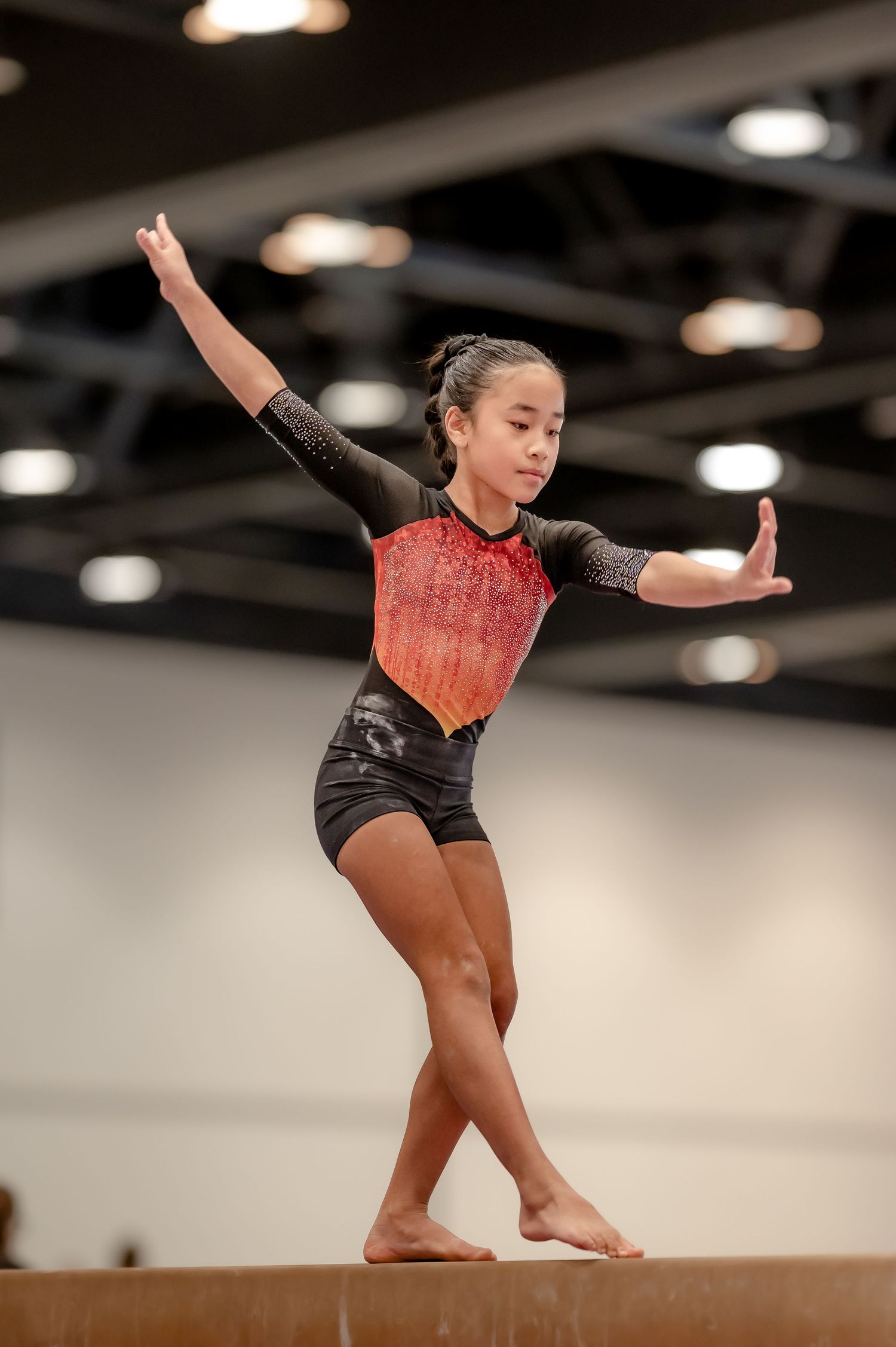 Young gymnast balancing on beam in black and red leotard with arms extended during competition.