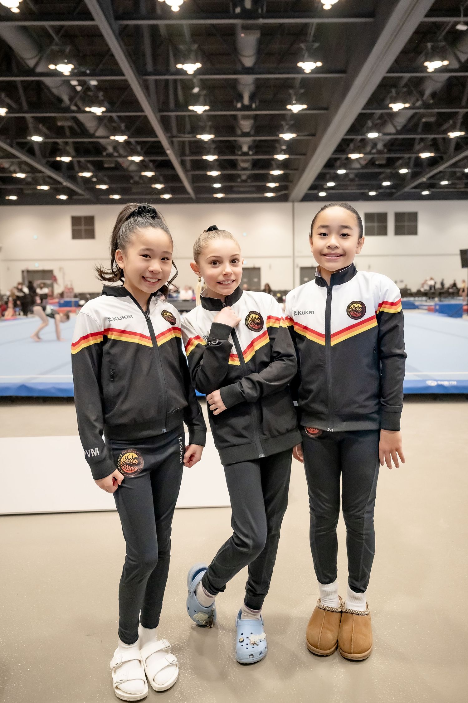 Three young gymnasts in matching black and red team jackets smile together at an indoor gymnastics competition.