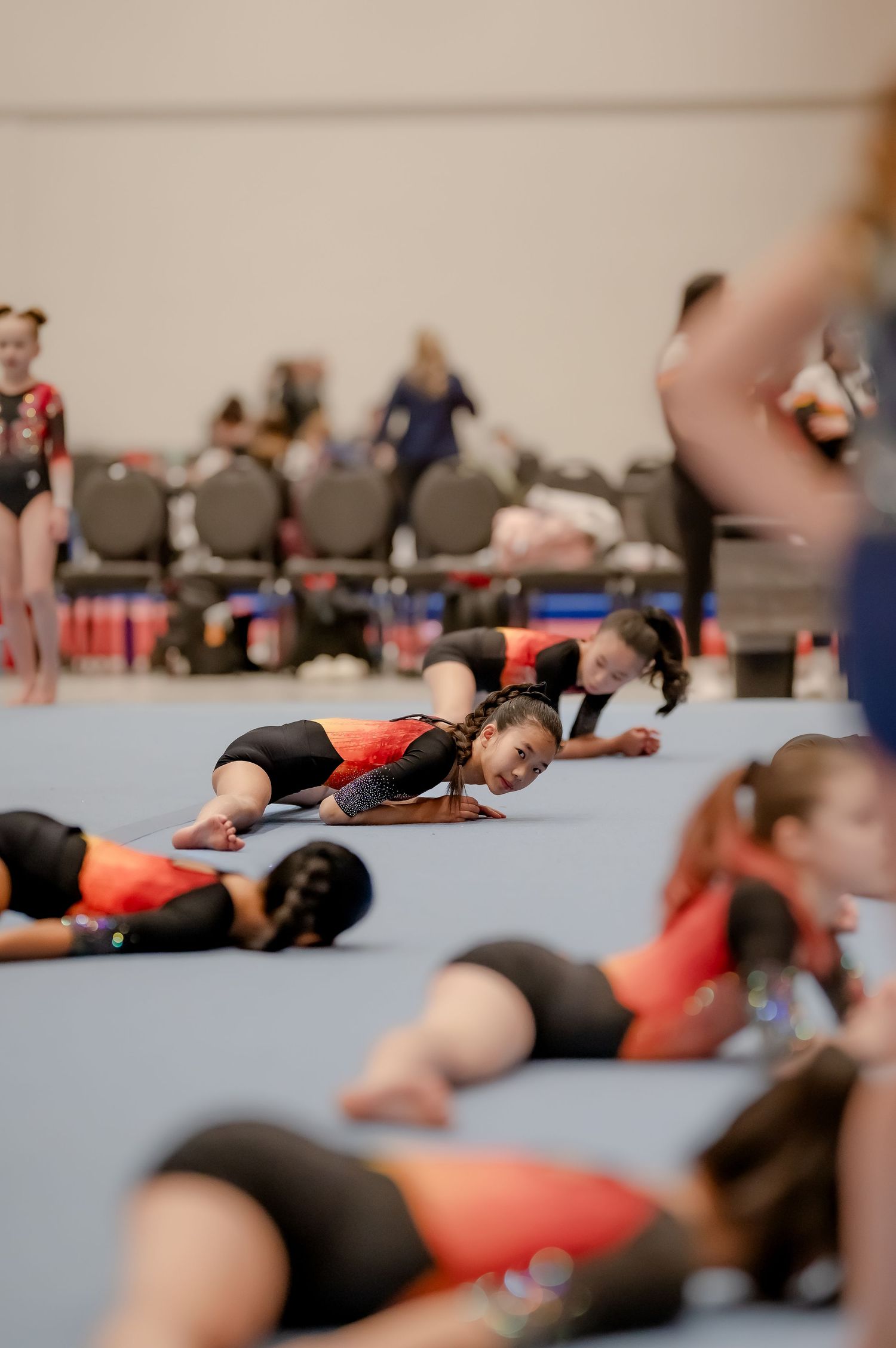 Young gymnasts in black and red leotards stretching on a blue floor mat during a gymnastics competition warmup.
