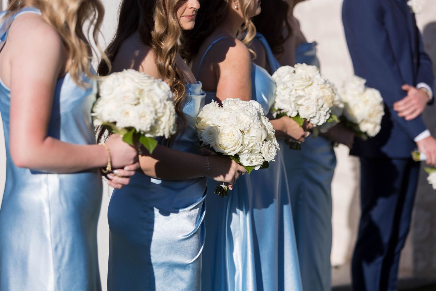 Bridesmaids in light blue satin dresses holding white hydrangea bouquets stand beside groomsmen in navy suits.